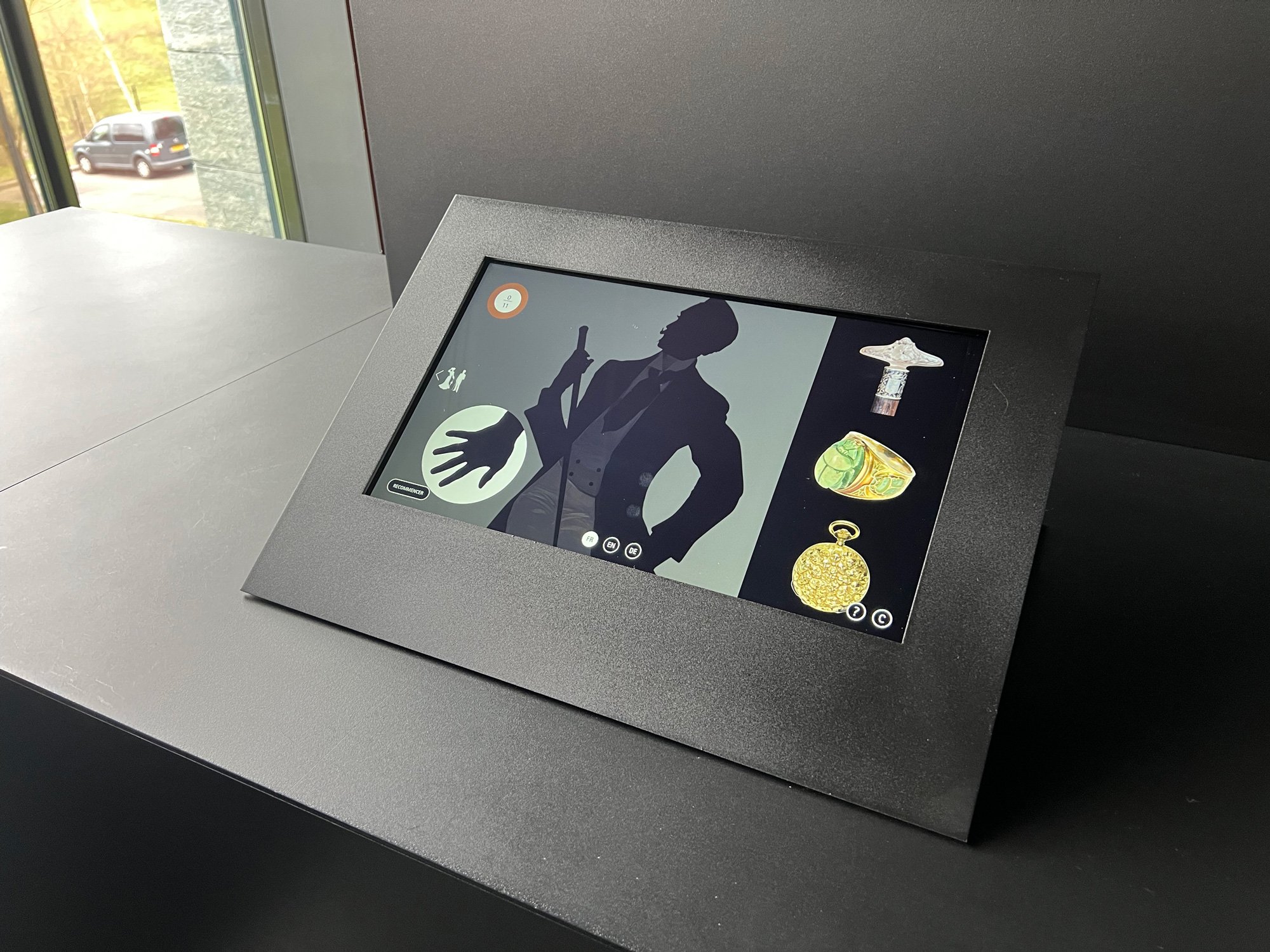 L'image montre un écran tactile placé sur une surface noire. L'écran présente des illustrations, dont une silhouette d'homme en costume avec une main posée, ainsi que plusieurs objets qui semblent être des artefacts ou bijoux, comme une montre de poche et une bague. Le fond de l'écran est sobre, mettant en valeur les éléments graphiques. On peut apercevoir à l'extérieur à travers une fenêtre, suggérant que l'image a été prise dans un espace d'exposition ou un musée.