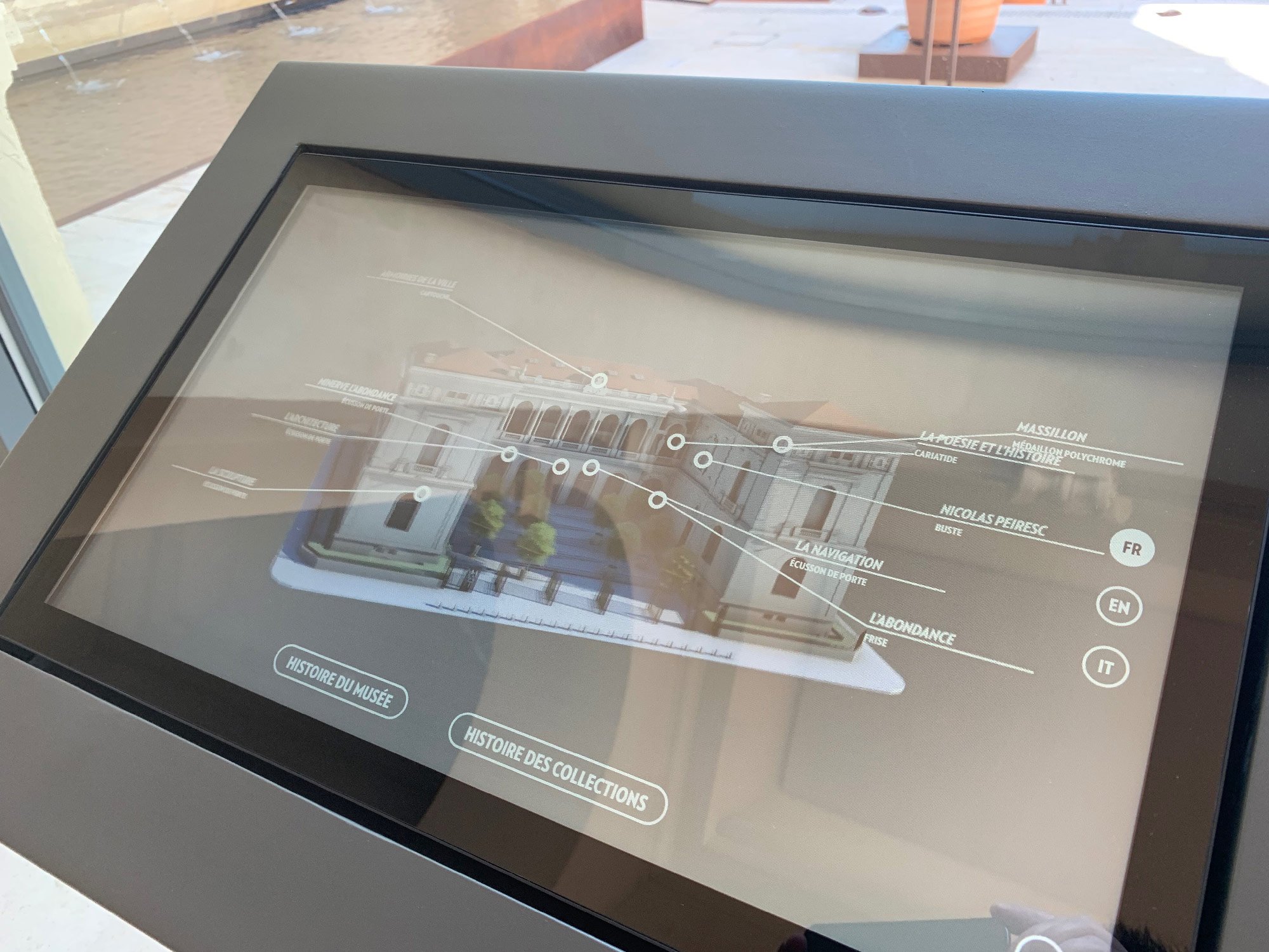 L'image montre un écran tactile interactif présentant une maquette ou une image en 3D d'un bâtiment, probablement un musée ou une galerie d'art. L'interface inclut des options de navigation en plusieurs langues, telles que le français (FR), l'anglais (EN) et l'italien (IT). Il y a des options pour explorer l'histoire du bâtiment ainsi que les collections qu'il abrite. En arrière-plan, on peut apercevoir un environnement extérieur, possiblement une terrasse ou un patio, avec de l'eau visible.