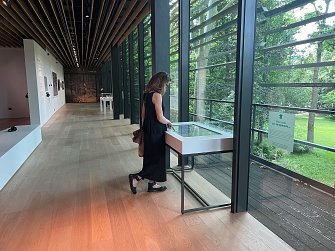 L'image montre une personne se tenant devant une vitrine dans un espace d'exposition. Elle examine quelque chose à l'intérieur de la vitrine, qui est éclairée par la lumière naturelle qui entre par de grandes fenêtres. On peut voir un paysage verdoyant à l'extérieur, suggérant que l'espace est entouré de nature. Le décor intérieur est moderne, avec des murs blancs et un sol en bois. La personne porte une robe noire et semble plongée dans sa contemplation.