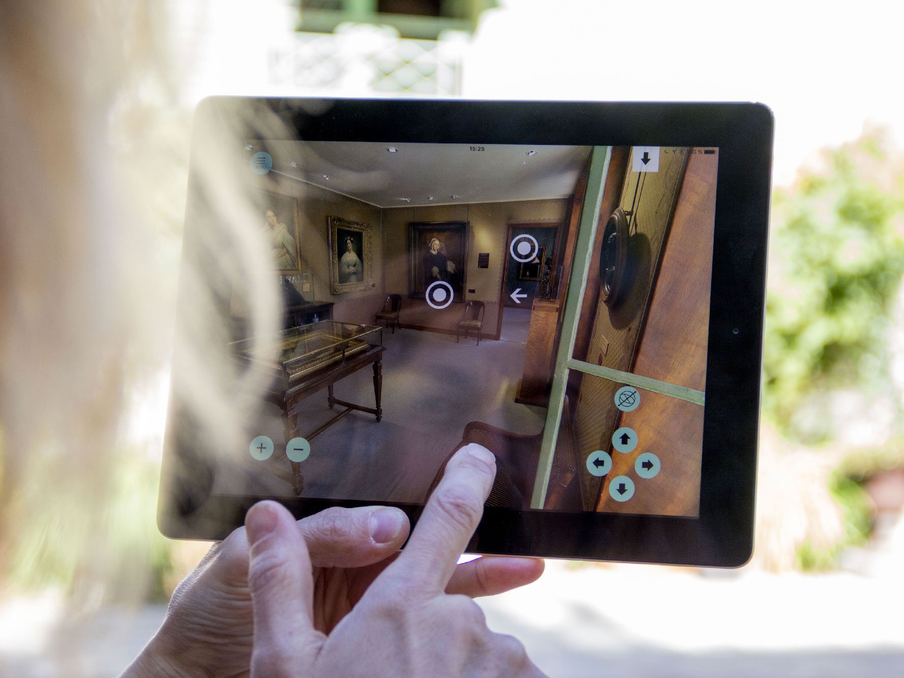 L'image montre une personne tenant une tablette dans un espace intérieur. Sur l'écran de la tablette, on peut voir une représentation en 3D d'une pièce, probablement issue d'une visite virtuelle. Des éléments de navigation, comme des cercles et des flèches, sont visibles sur l'écran, suggérant que l'utilisateur peut interagir avec l'environnement virtuel. L'arrière-plan de la photo montre une zone bien éclairée, probablement un jardin ou un patio, ce qui donne une ambiance agréable à la scène.
