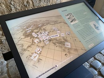 L'image montre un écran interactif qui présente une carte de la région de l'Océanie. Plusieurs petites icônes ou images sont dispersées sur la carte, probablement pour illustrer des points d'intérêt ou des événements historiques. À droite de l'écran, il y a un texte explicatif qui pourrait fournir des informations sur l'exploration géographique et maritime de cette région. L'ensemble est installé dans un environnement qui semble être un musée ou un espace d'information historique.