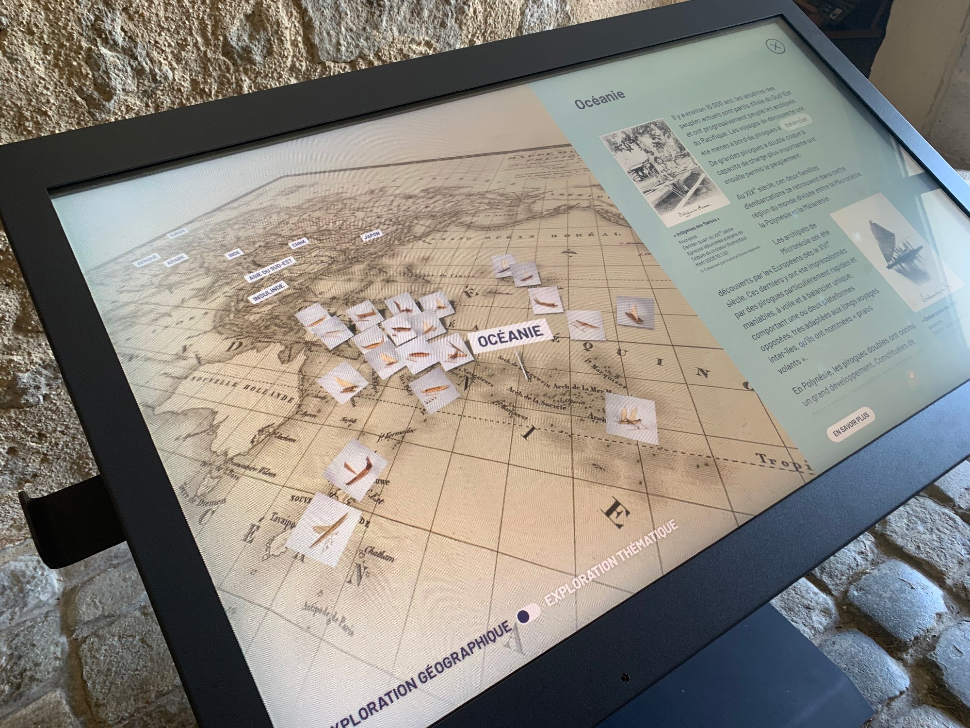 L'image montre un écran interactif qui présente une carte de la région de l'Océanie. Plusieurs petites icônes ou images sont dispersées sur la carte, probablement pour illustrer des points d'intérêt ou des événements historiques. À droite de l'écran, il y a un texte explicatif qui pourrait fournir des informations sur l'exploration géographique et maritime de cette région. L'ensemble est installé dans un environnement qui semble être un musée ou un espace d'information historique.