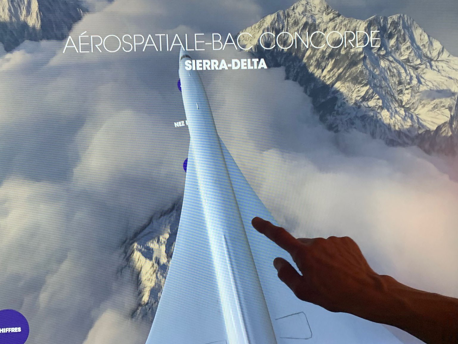 L'image montre une interface numérique affichant des informations sur l'aéronautique, plus particulièrement sur le Concorde, avec le mot "SIERRA-DELTA" en évidence. Au premier plan, une main touche l'écran, indiquant une interaction avec l'affichage. En arrière-plan, on aperçoit un paysage montagneux couvert de nuages, ajoutant une dimension visuelle impressionnante à l'ensemble. Des options de langue (FR, EN) sont également présentes en bas de l'image.