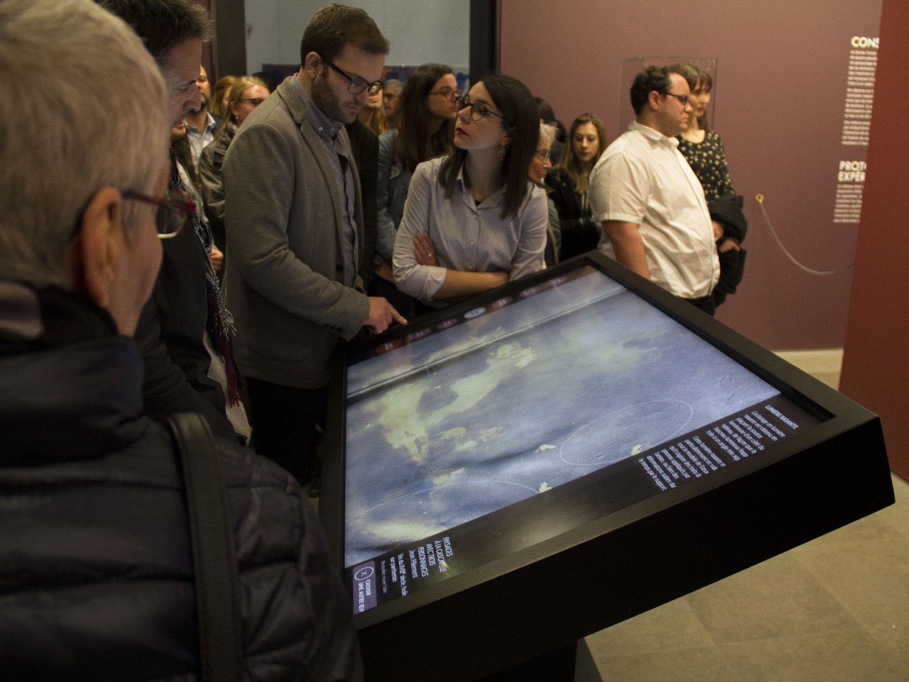 L'image montre un groupe de personnes rassemblées autour d'un grand écran tactile interactif. Deux personnes, probablement un homme et une femme, semblent particulièrement intéressées par ce qui est affiché à l'écran. L'arrière-plan est flou, mais on peut voir d'autres visiteurs curieux observant l'exposition. Le décor semble être celui d'un musée ou d'une galerie, avec des murs d'une couleur sombre.
