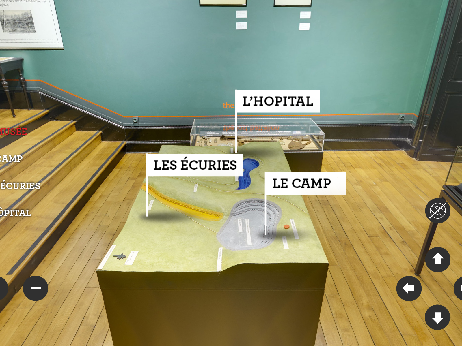 L'image montre une exposition dans un musée. Sur une table d'exposition, plusieurs éléments de présentation sont étiquetés. On y trouve les termes « l'Hôpital », « les Écuries » et « le Camp ». L'environnement est élégant, avec des murs de couleur verte et un sol en bois. Des objets ou maquettes semblent être présentés sous une vitrine, suggérant une thématique historique.