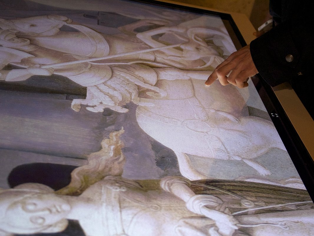 L'image montre une personne pointant du doigt une œuvre d'art affichée sur un écran. L'œuvre représente une scène détaillée, probablement historique ou religieuse, avec des figures équestres et des personnages. Les couleurs sont délicates et les détails sont fins, suggérant un style classique. La personne interagit avec l'image, attirant l'attention sur un détail spécifique de la composition.