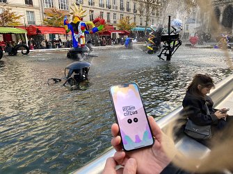 L'image montre une scène animée dans un espace public, probablement un espace culturel ou artistique. Au premier plan, une personne tient un téléphone portable sur lequel on peut voir un écran affichant le mot "ircam circus". En arrière-plan, il y a une grande fontaine avec de l'eau qui s'écoule, entourée de différentes sculptures colorées et ludiques. À droite, une autre personne est assise, semblant contempler la scène. Les arbres sont visibles, et l'atmosphère paraît vivante grâce aux couleurs vives des sculptures et aux éléments interactifs de la fontaine.