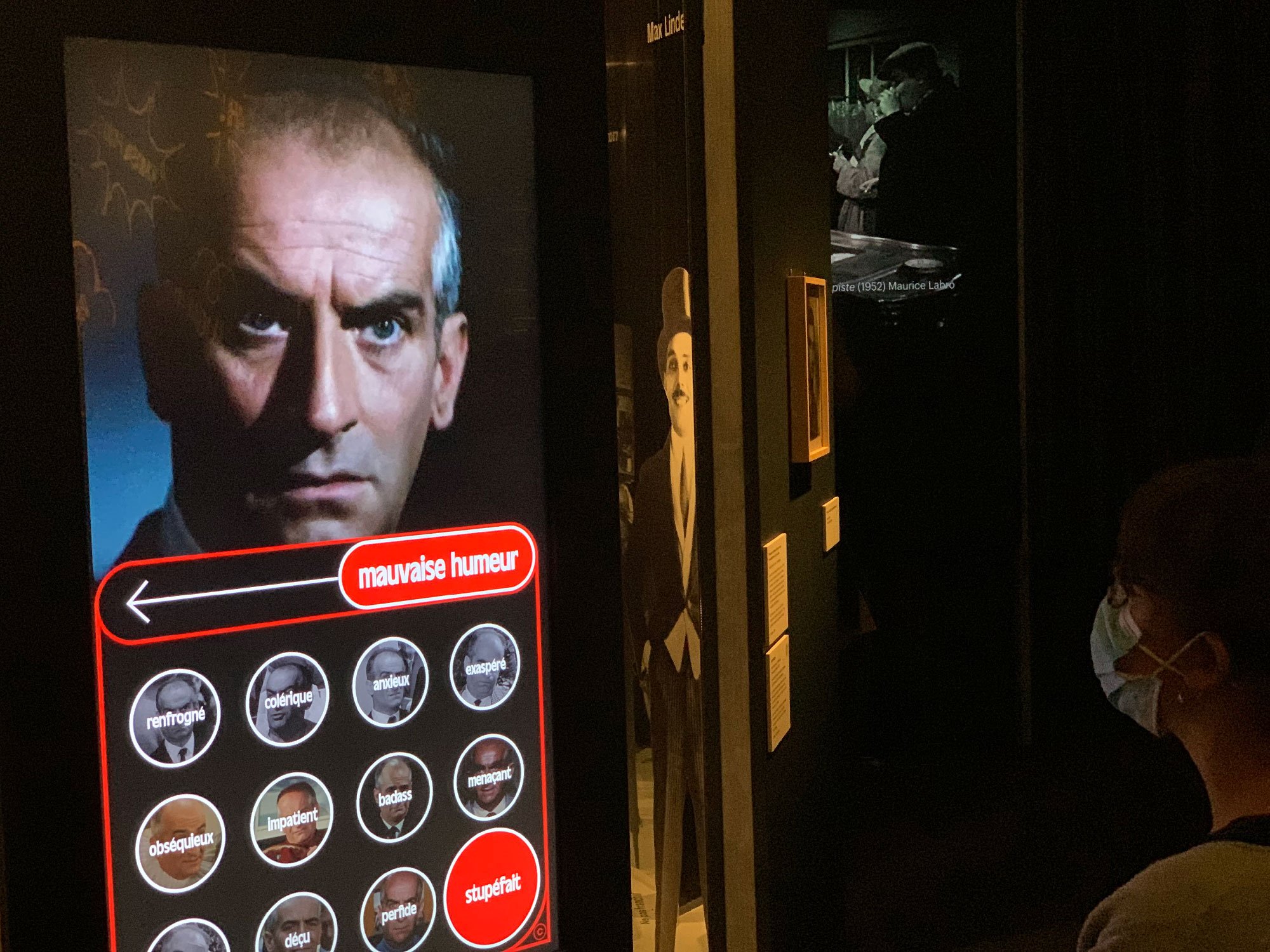 L'image montre un écran interactif d'une exposition où une personne semble interagir avec une interface. À l'écran, on voit un portrait d'un homme avec une expression sérieuse, accompagnée de choix d'émotions comme "mauvaise humeur", "stressé", et d'autres. Au fond, on aperçoit des affiches ou des images en noir et blanc, probablement liées au thème de l'exposition. La personne devant l'écran porte un masque, indiquant que l'environnement est propice à des mesures sanitaires.