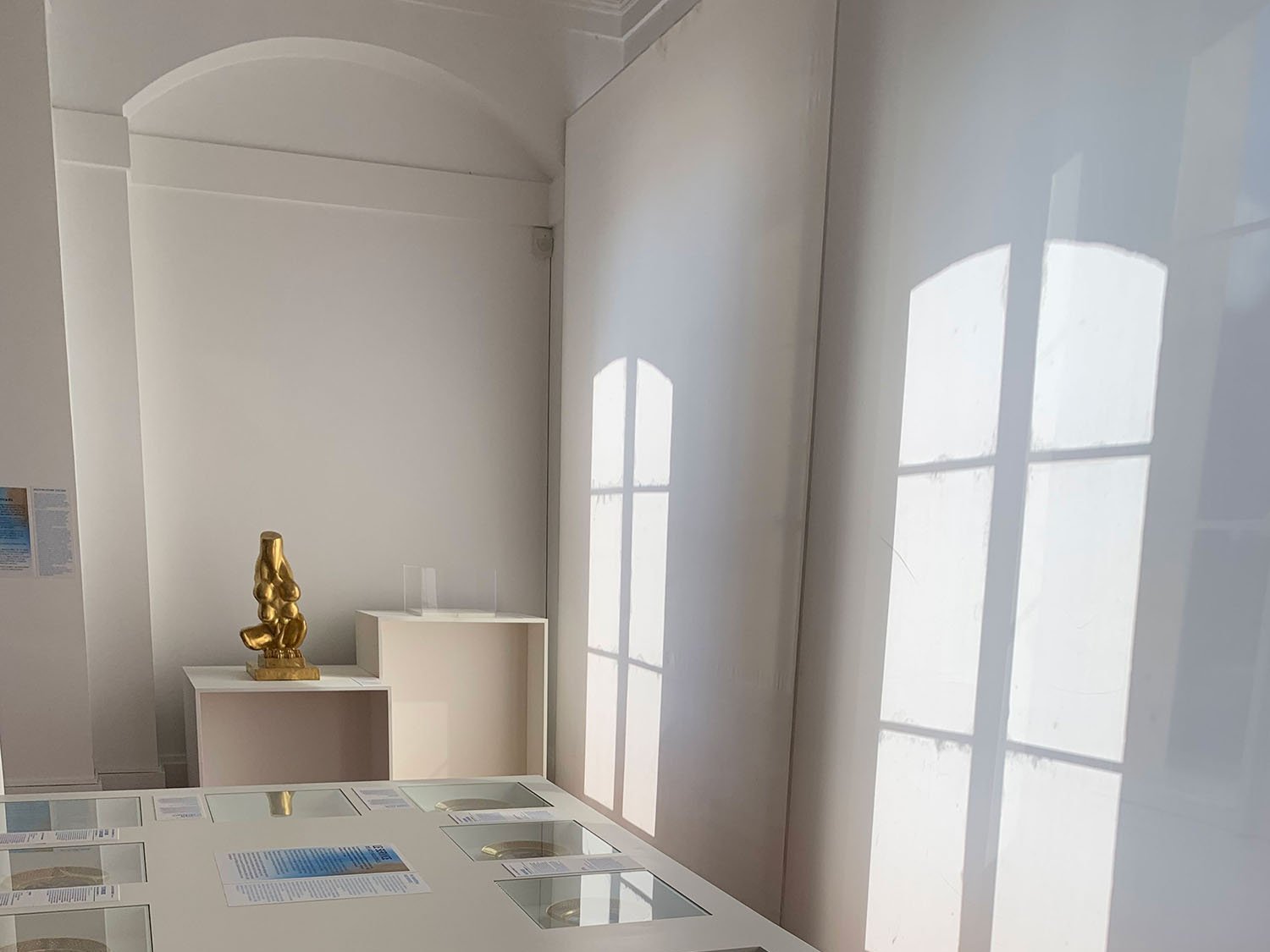 L'image montre une salle d'exposition avec un éclairage naturel provenant de fenêtres. Au centre, il y a une table blanche avec un écran tactile qui présente divers éléments visuels. Autour de cette table se trouvent des cadres avec des images ou des informations. À l'arrière-plan, il y a un objet doré sur une autre table, et l'ensemble de l'espace dégage une ambiance épurée et moderne grâce aux couleurs claires et au parquet en bois. Les ombres créées par la lumière filtrée ajoutent une touche esthétique à la scène.