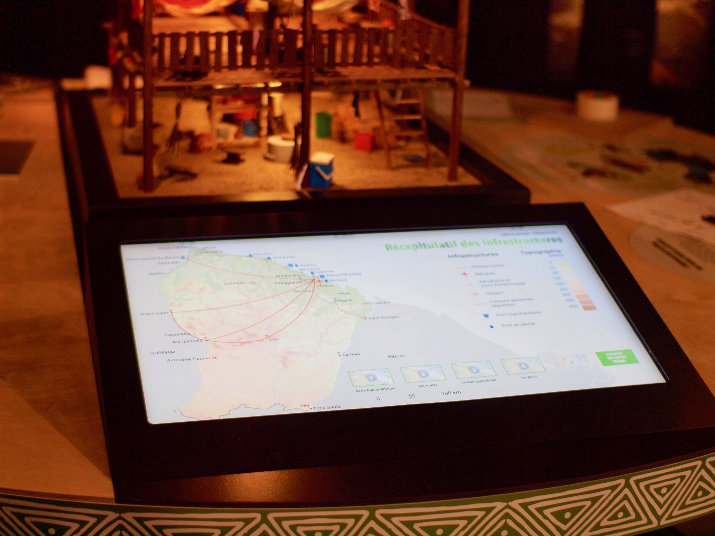 L'image montre un écran tactile présentant une carte avec des informations sur les infrastructures. On peut voir des lignes reliant différentes localités, probablement pour illustrer des connexions ou des réseaux. En arrière-plan, il y a une maquette d'une structure, suggérant qu'il s'agit d'une exposition ou d'un musée. Les éléments colorés sur la maquette évoquent des activités ou des espaces de vie. L'écran affiche également des icônes et des légendes, indiquant des données supplémentaires ou des statistiques.