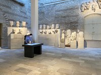 L'image montre un espace intérieur, probablement un musée ou une galerie. On peut y voir plusieurs sculptures en marbre, dont des bustes figurants des visages humains, alignés sur des socles le long des murs. Au centre, une personne se tient à un bureau noir, regardant attentivement quelque chose sur la surface. La lumière naturelle éclaire la pièce, créant une atmosphère calme et contemplative. Les murs en pierre et le sol en dalles ajoutent une note historique à l'environnement.