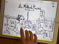 L'image montre une illustration en noir et blanc affichée sur un écran d'iPad. On y voit une femme, probablement inspirée de Marie Curie, tenant des éprouvettes dans un laboratoire. En arrière-plan, il y a plusieurs instruments scientifiques, ainsi qu'un portrait représentant des personnages, qui semblent être liés à l'histoire de Marie Curie. Un titre en haut de l'image indique "La Méthode Curie". Une main touche l'écran, indiquant une interaction avec l'illustration.