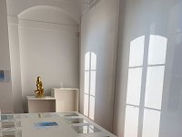 L'image montre une salle d'exposition avec un éclairage naturel provenant de fenêtres. Au centre, il y a une table blanche avec un écran tactile qui présente divers éléments visuels. Autour de cette table se trouvent des cadres avec des images ou des informations. À l'arrière-plan, il y a un objet doré sur une autre table, et l'ensemble de l'espace dégage une ambiance épurée et moderne grâce aux couleurs claires et au parquet en bois. Les ombres créées par la lumière filtrée ajoutent une touche esthétique à la scène.