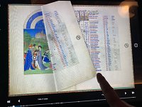 L'image montre un folio d'un manuscrit ancien, probablement un livre d'heures ou un autre texte manuscrit. Sur la gauche, il y a une illustration colorée représentant un groupe de personnages en costumes médiévaux, probablement dans un décor de château ou de paysage. À droite, il y a une liste de noms ou de mots écrits dans une calligraphie soignée. Un doigt pointe vers la page, suggérant que la personne montre un détail particulier de l'illustration ou du texte. Le tout est encadré d'une bordure décorative qui ajoute à l'élégance du manuscrit.