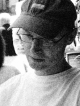 L'image montre un homme portant une casquette et des lunettes. Il est concentré sur quelque chose qu'il lit ou observe, le regard baissé. L'arrière-plan présente des silhouettes floues d'autres personnes, suggérant une atmosphère sociale. Le contraste entre les tons clairs et sombres donne un effet artistique à la photo.
