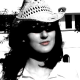 L'image montre une femme portant un chapeau de cow-boy en paille. Elle a des cheveux longs et sombres et affiche une expression confiante. La photo est en noir et blanc, ce qui accentue les contrastes et donne une ambiance nostalgique. En arrière-plan, on devine des éléments architecturaux, mais ils sont flous.