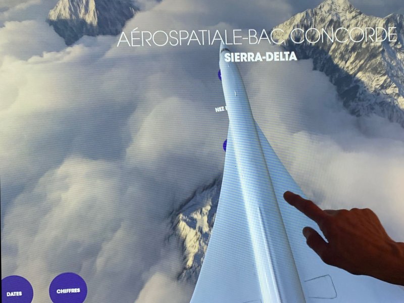 L'image montre une interface numérique affichant des informations sur l'aéronautique, plus particulièrement sur le Concorde, avec le mot "SIERRA-DELTA" en évidence. Au premier plan, une main touche l'écran, indiquant une interaction avec l'affichage. En arrière-plan, on aperçoit un paysage montagneux couvert de nuages, ajoutant une dimension visuelle impressionnante à l'ensemble. Des options de langue (FR, EN) sont également présentes en bas de l'image.