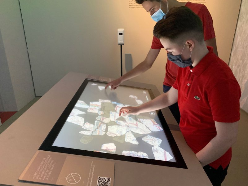 L'image montre deux jeunes garçons interactant avec un écran tactile en exposant des éléments. Ils portent des masques en raison de mesures sanitaires. L'écran affiche de nombreux morceaux colorés, peut-être des artefacts ou des éléments informatiques, et les garçons semblent manipuler ces objets virtuels. L'environnement est moderne et éducatif, suggérant qu'ils se trouvent dans un musée ou une exposition.