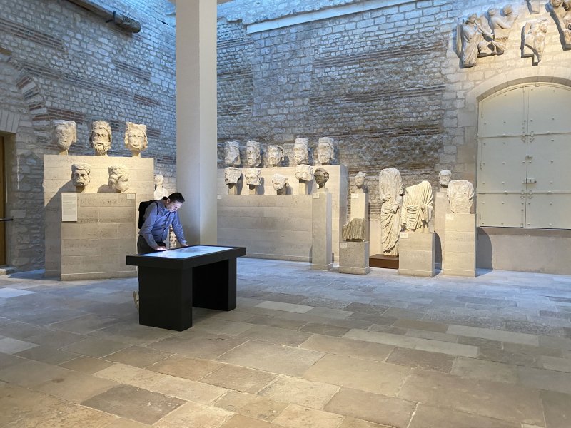 L'image montre un espace intérieur, probablement un musée ou une galerie. On peut y voir plusieurs sculptures en marbre, dont des bustes figurants des visages humains, alignés sur des socles le long des murs. Au centre, une personne se tient à un bureau noir, regardant attentivement quelque chose sur la surface. La lumière naturelle éclaire la pièce, créant une atmosphère calme et contemplative. Les murs en pierre et le sol en dalles ajoutent une note historique à l'environnement.